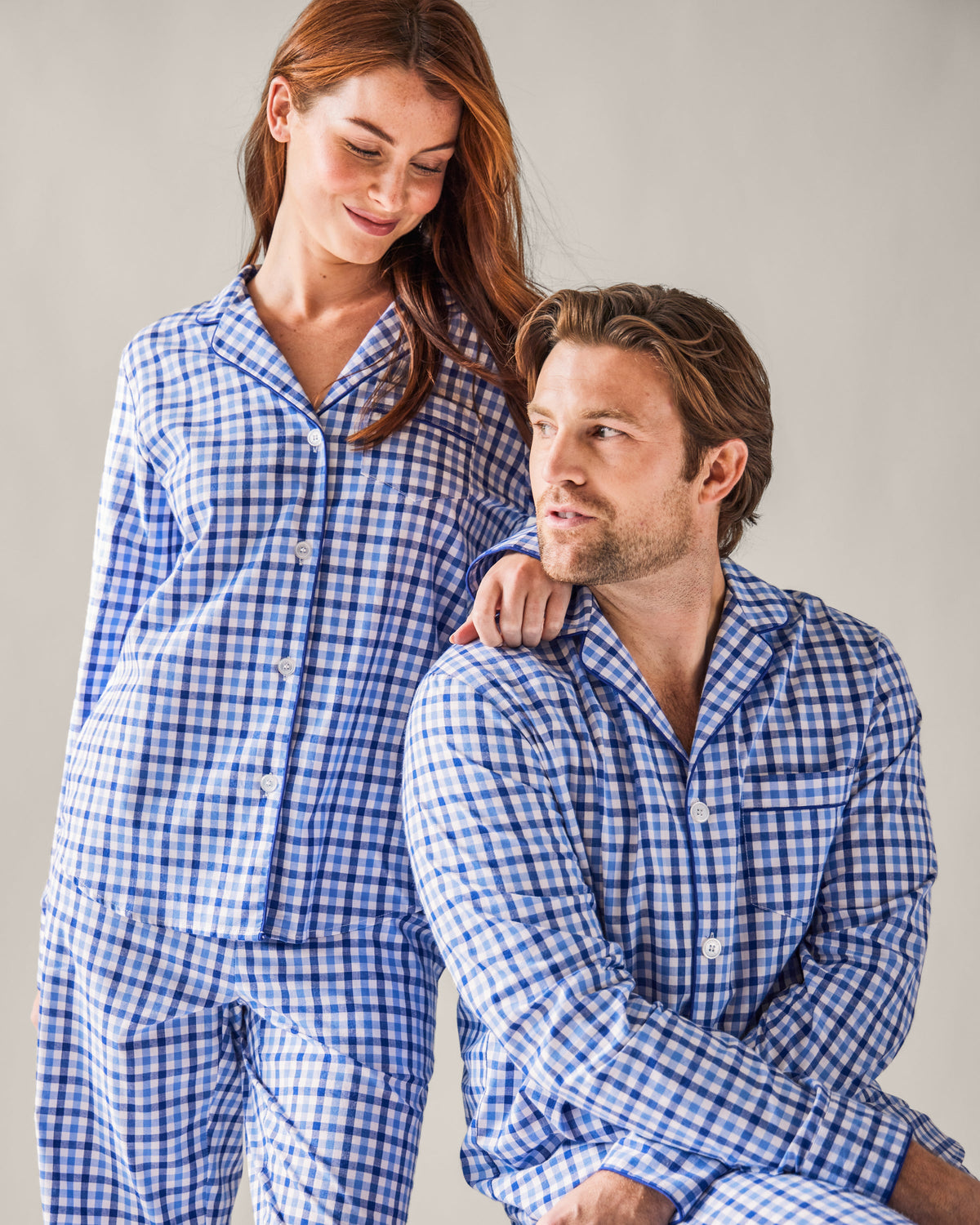 A man and woman wear matching Petite Plume pajamas. The man, seated, wears the Mens Twill Pajama Set in Royal Blue Gingham while the woman stands with her hand on his shoulder. They smile at each other against a plain background, highlighting their fine cotton attire.