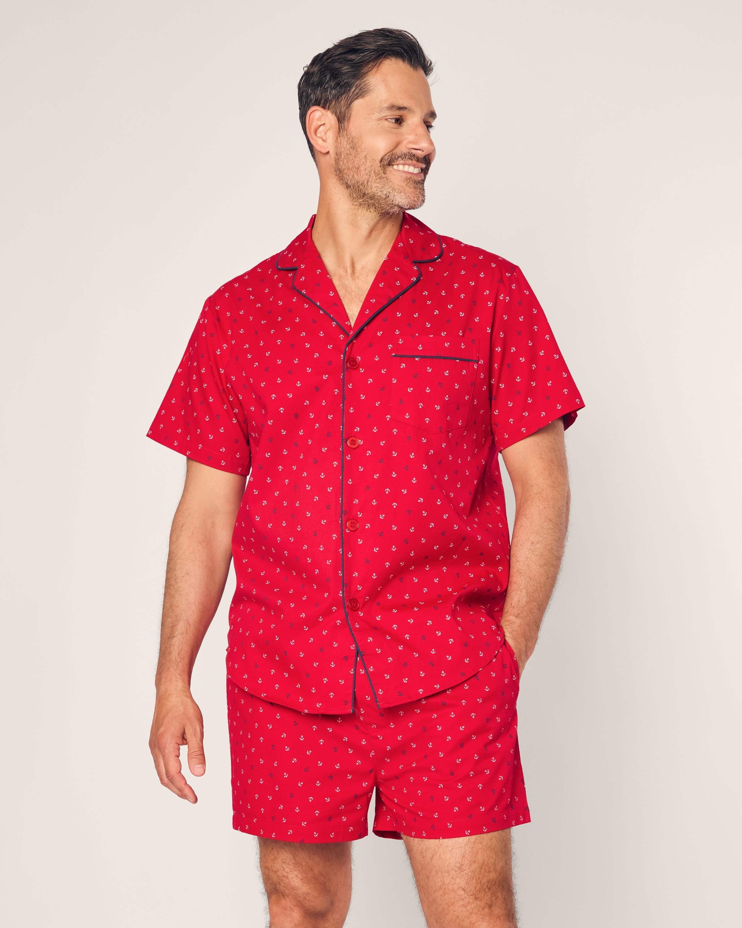 A man smiles and looks right, clad in a Petite Plume Mens Twill Short Sleeve Short Set Anchors Away in matching red. Made from premium cotton, he rests his left hand in his pocket against a plain, light-colored background.