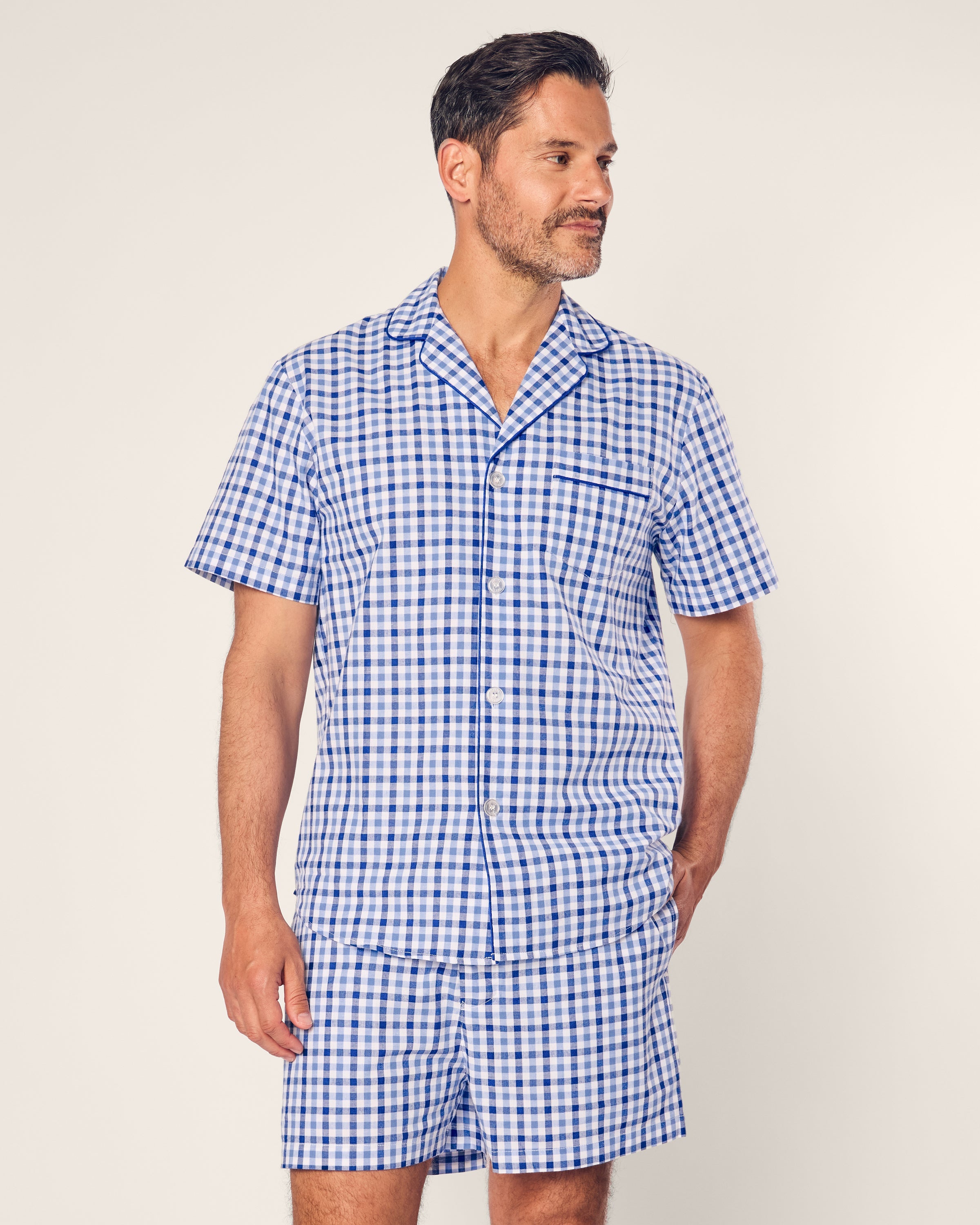 A bearded man with short hair, clad in a Mens Twill Pajama Short Set by Petite Plume in Royal Blue Gingham, gazes to the side, exuding comfort against a plain light gray background.