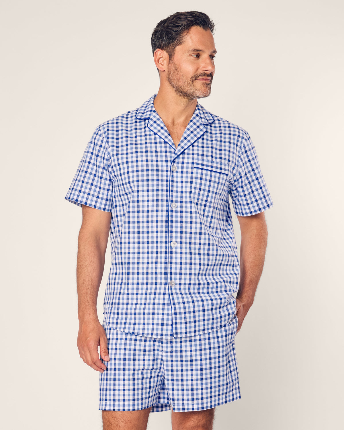 A bearded man with short hair, clad in a Mens Twill Pajama Short Set by Petite Plume in Royal Blue Gingham, gazes to the side, exuding comfort against a plain light gray background.