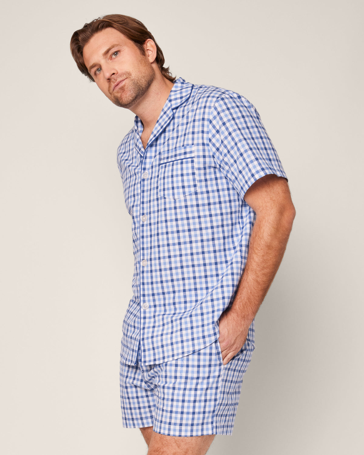 A man wearing the Petite Plume Mens Twill Pajama Short Set in Royal Blue Gingham tilts slightly to the side against a plain, light-colored background, highlighting the luxury sleepwear crafted from the finest quality cotton.