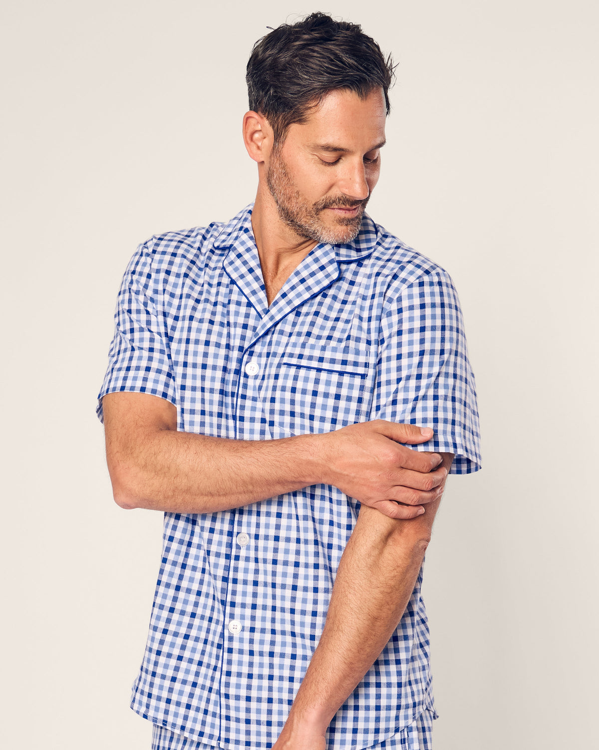 A man in the Petite Plume Mens Twill Pajama Short Set, featuring a blue and white gingham pattern, crafted from premium cotton, touches his left arm with his right hand against a plain light background.