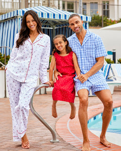 A woman in nautical print cotton pajamas, a child in a red polka-dot dress, and a man in Petite Plumes Mens Twill Pajama Short Set in Royal Blue Gingham smile by the pool with a charming striped cabana backdrop.