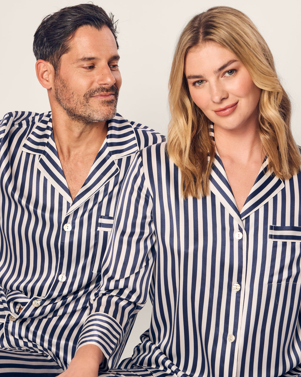 A man and woman sit side by side in matching Petite Plume Mens Silk Pajama Sets in Navy Bengal Stripe. The man, with short dark hair and a beard, smiles at the blonde-haired woman against a plain backdrop—embodying luxury sleepwear comfort.