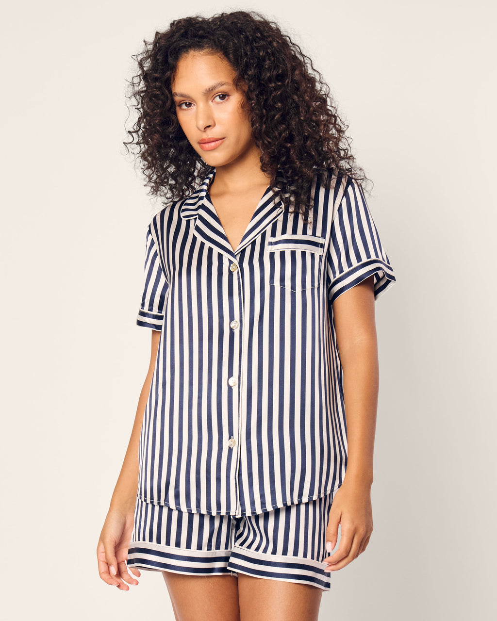 A woman with curly hair stylishly wears the Womens Silk Short Set in Navy Bengal Stripe by Petite Plume. This luxury sleepwear features short sleeves and shorts in navy blue and white stripes on a light background.
