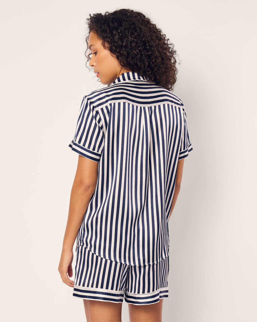 A person with curly hair stands facing away, dressed in Petite Plumes Womens Silk Short Set in Navy Bengal Stripe. The matching shirt and shorts exude luxury sleepwear elegance against a plain, light backdrop.