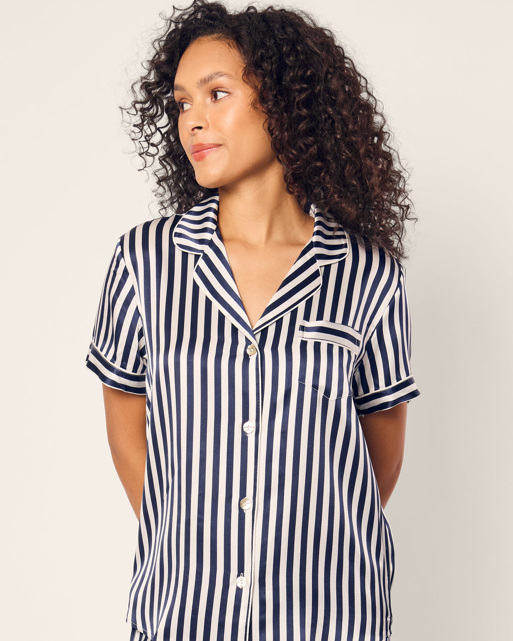 A curly-haired individual wears the Womens Silk Short Set in Navy Bengal Stripe by Petite Plume, gazing to the side against a neutral background.