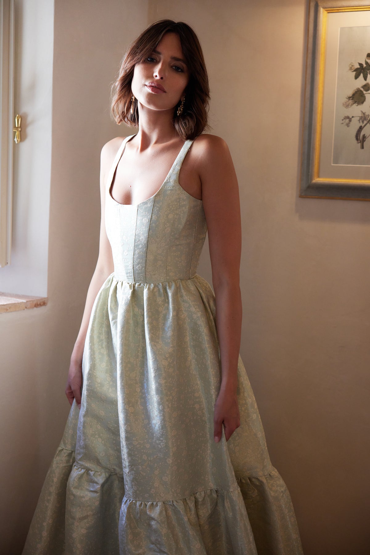 Front view of The Avery Dress in Sage Duchess showcasing its scoop neckline, corseted bodice, floral details, and gathered tiers indoors