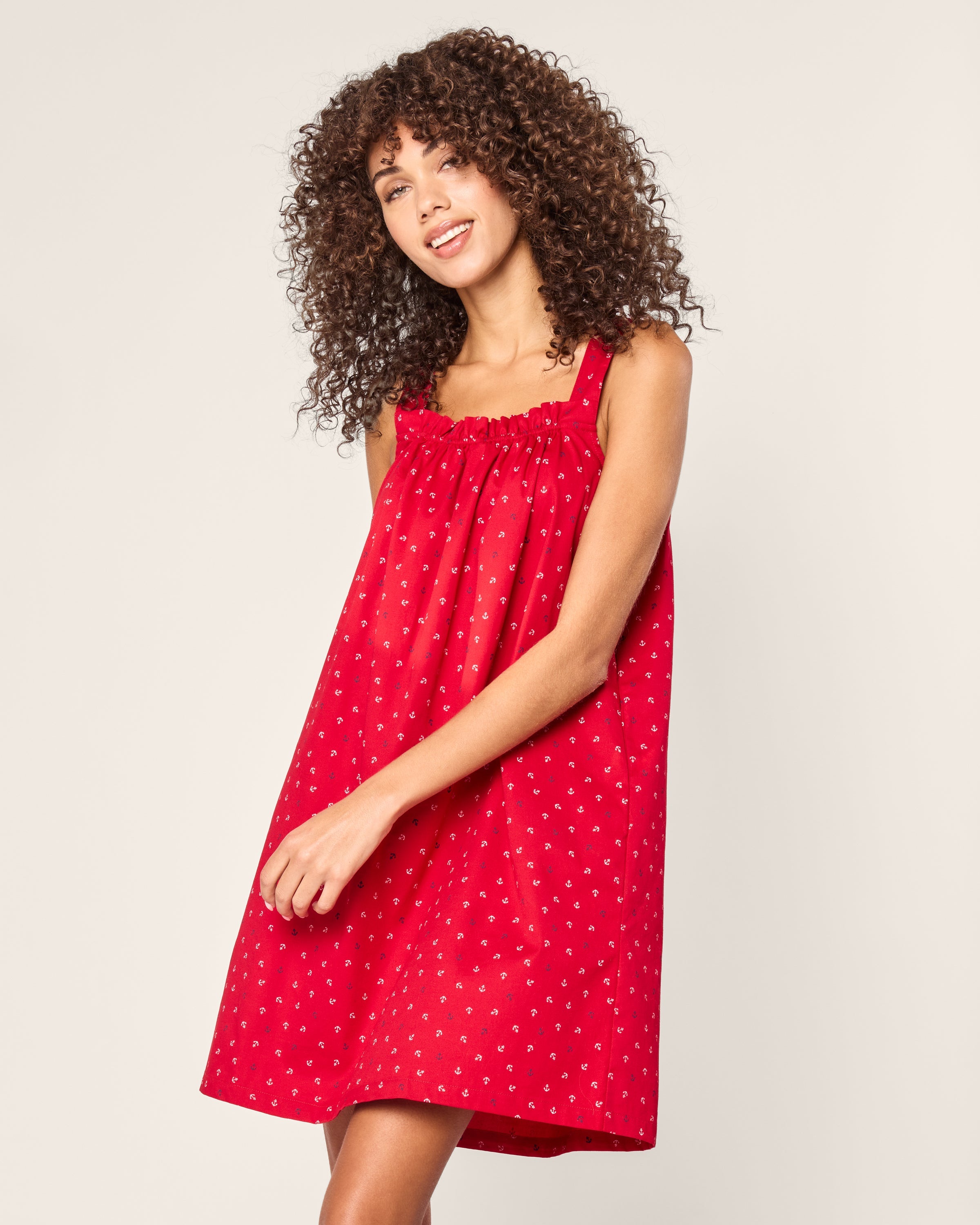 A person with curly hair in a red sleeveless Petite Plume Womens Twill Annabelle Nightgown, featuring a delicate anchors print, stands against a plain background. They smile and glance aside, exuding an effortless elegance reminiscent of luxury cotton sleepwear.