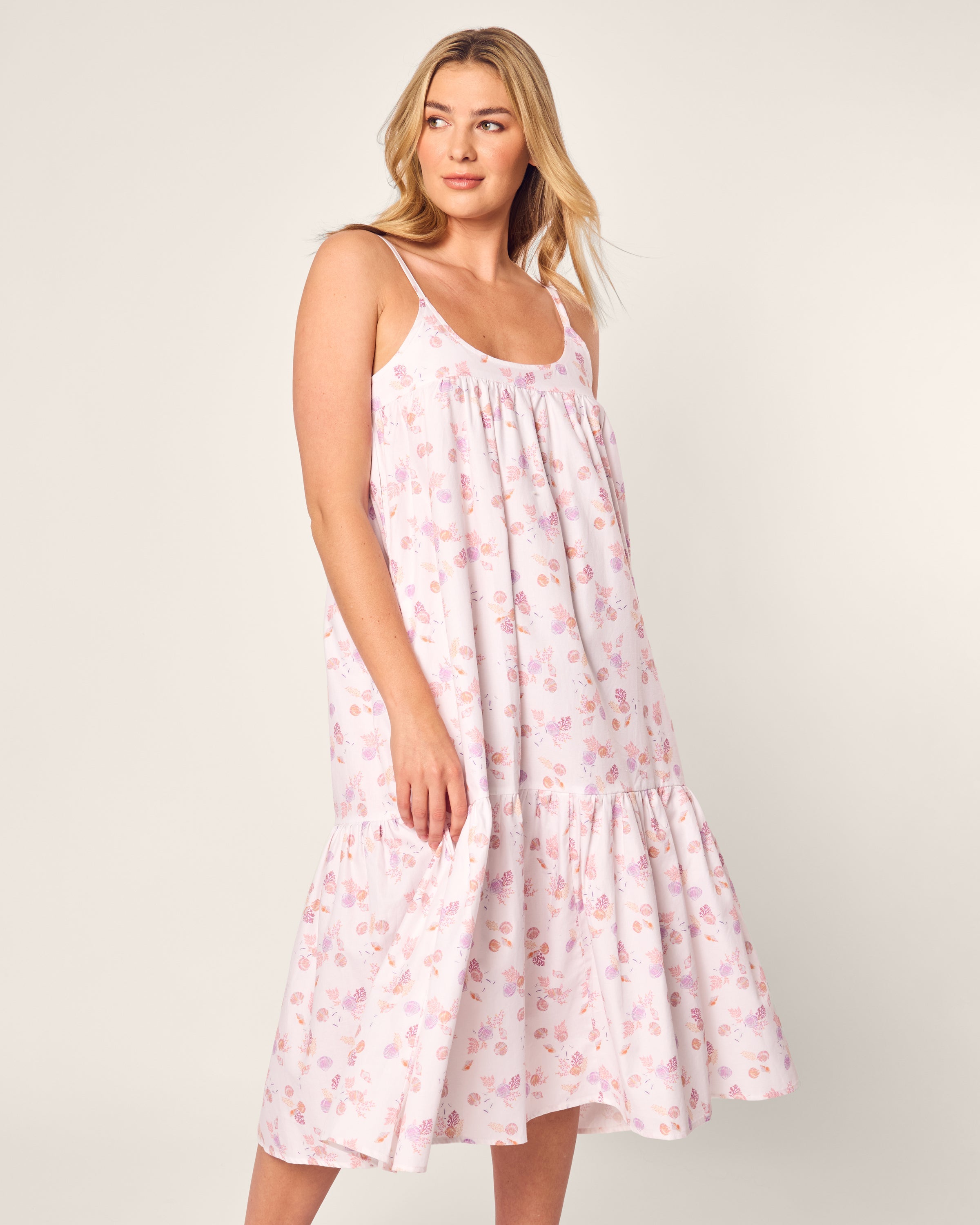 Dressed in the Womens Twill Chloe Nightgown by Petite Plume in Seashell Splendor, a person is captured against a plain backdrop, gazing sideways with a soft smile. The sleeveless white nightgown displays a floral pattern and ruffled hem, evoking luxury sleepwears flowy elegance.