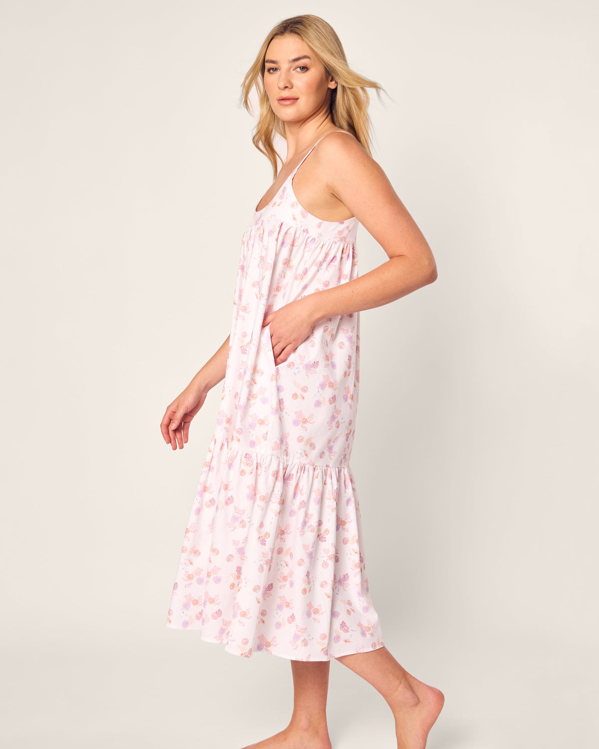 A person with long blond hair is wearing the Womens Twill Chloe Nightgown in Seashell Splendor by Petite Plume. This sleeveless, light pink nightgown has a floral pattern and soft cotton fabric, offering a flowing, relaxed fit. They stand barefoot against a plain background.