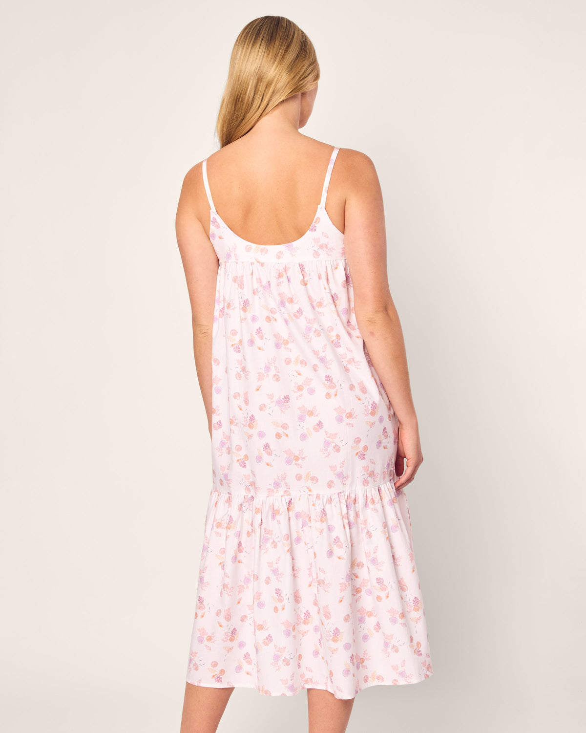 A person with long blonde hair is wearing the sleeveless Womens Twill Chloe Nightgown in Seashell Splendor by Petite Plume. Crafted from soft cotton, it features a pink floral design and a loose fit, reaching below the knees as the person faces away from the camera against a plain background.
