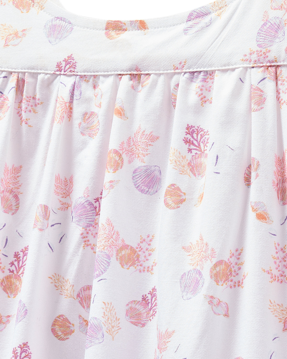 Close-up of Petite Plumes Womens Twill Chloe Nightgown in Seashell Splendor, featuring a white cotton fabric with pink and purple seashells and coral patterns. The luxury sleepwear displays gentle folds and has a soft texture.