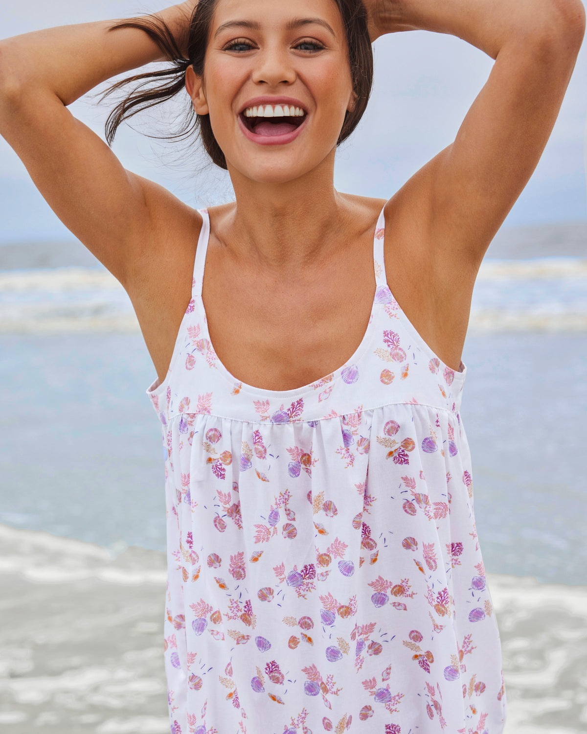 A woman in Petite Plumes Womens Twill Chloe Nightgown in Seashell Splendor smiles with her hands behind her head on a breezy beach, enjoying the serenity of the waves.