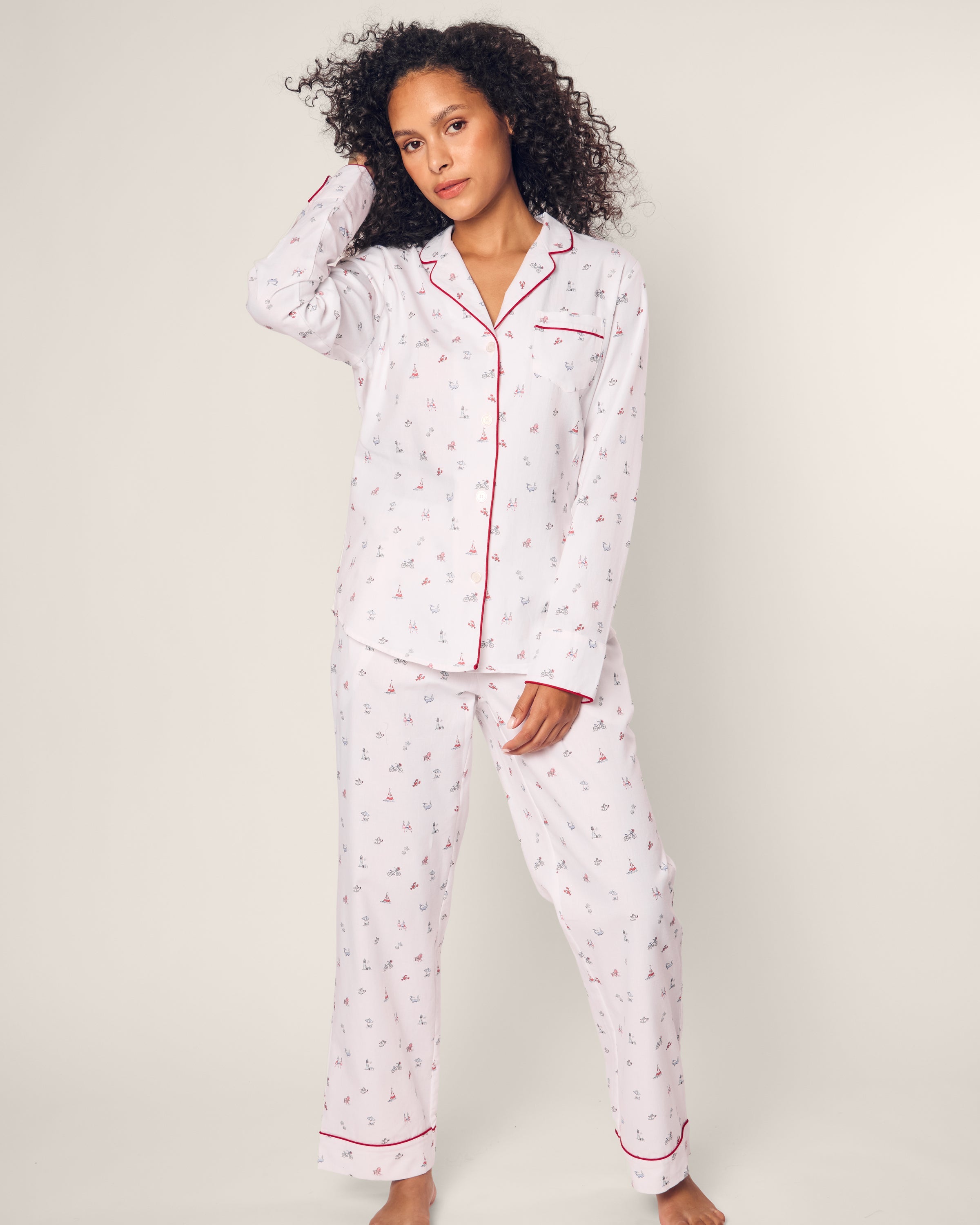 A person with curly hair wears the Petite Plume Womens Twill Pajama Set in Coastal Charms. This luxurious white cotton set features small red and blue patterns and elegant red piping. They stand barefoot, one hand touching their head, against a neutral background, exuding charm.