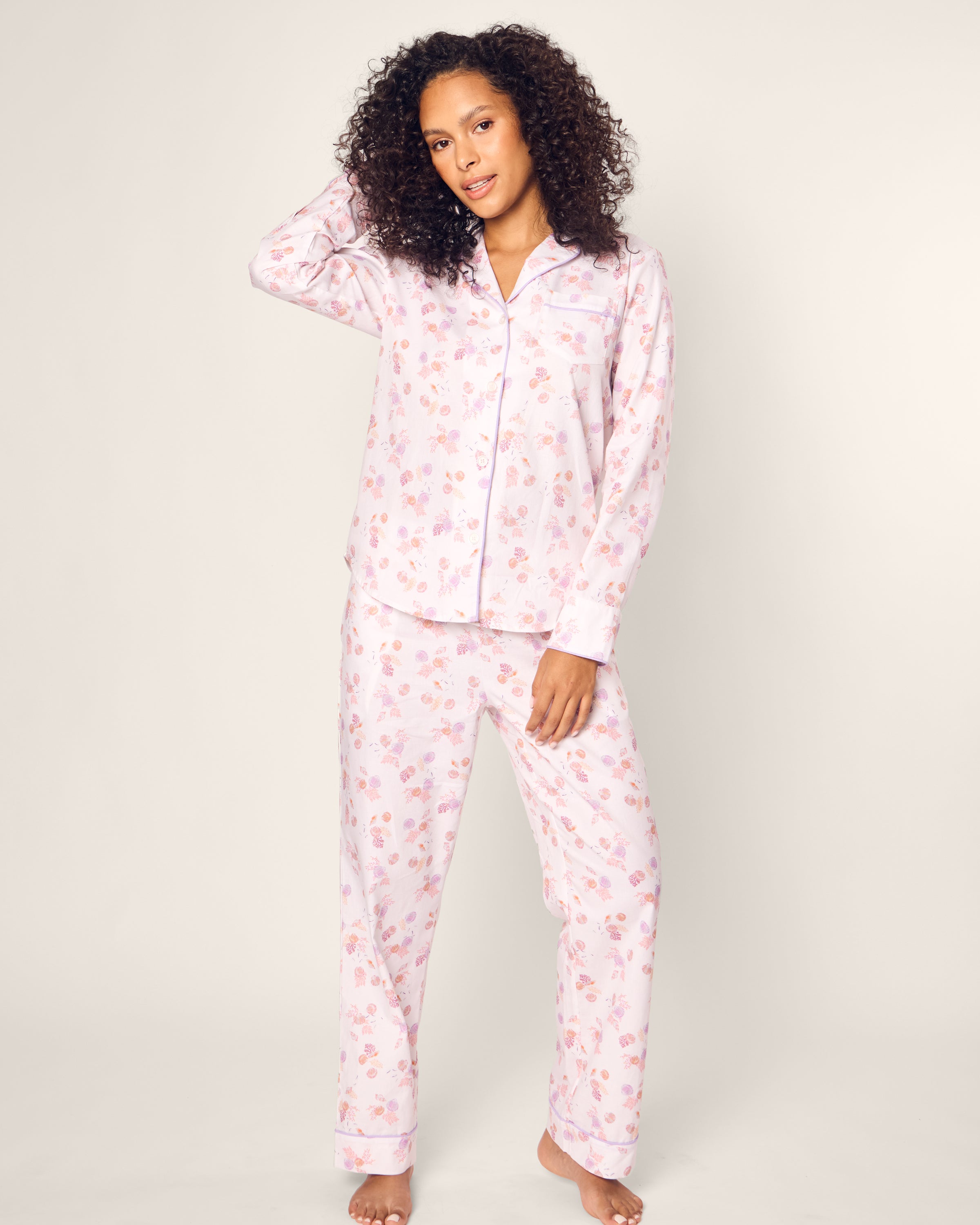 Standing barefoot on a white background, a person models Petite Plumes Womens Twill Pajama Set in Seashell Splendor. Featuring a long-sleeve shirt and pants, this luxurious ensemble ensures ultimate comfort and style as they touch their curly hair and gaze at the camera.