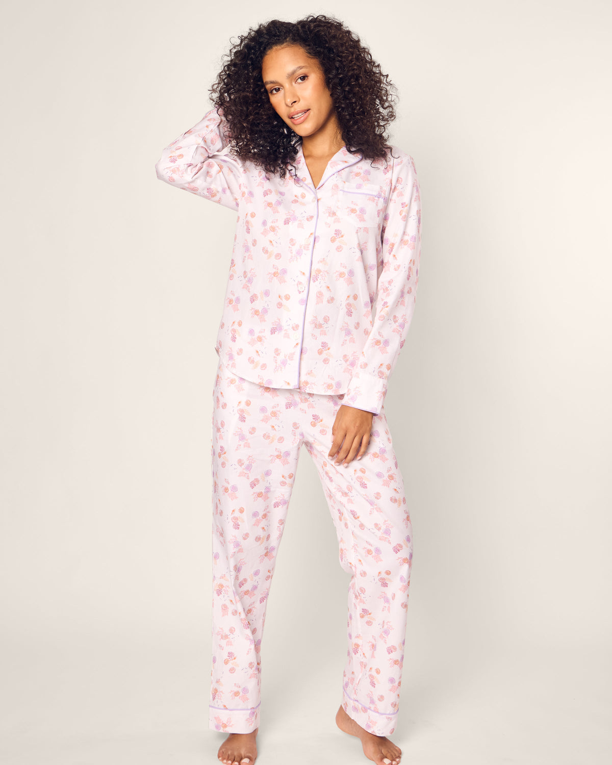 Standing barefoot on a white background, a person models Petite Plumes Womens Twill Pajama Set in Seashell Splendor. Featuring a long-sleeve shirt and pants, this luxurious ensemble ensures ultimate comfort and style as they touch their curly hair and gaze at the camera.