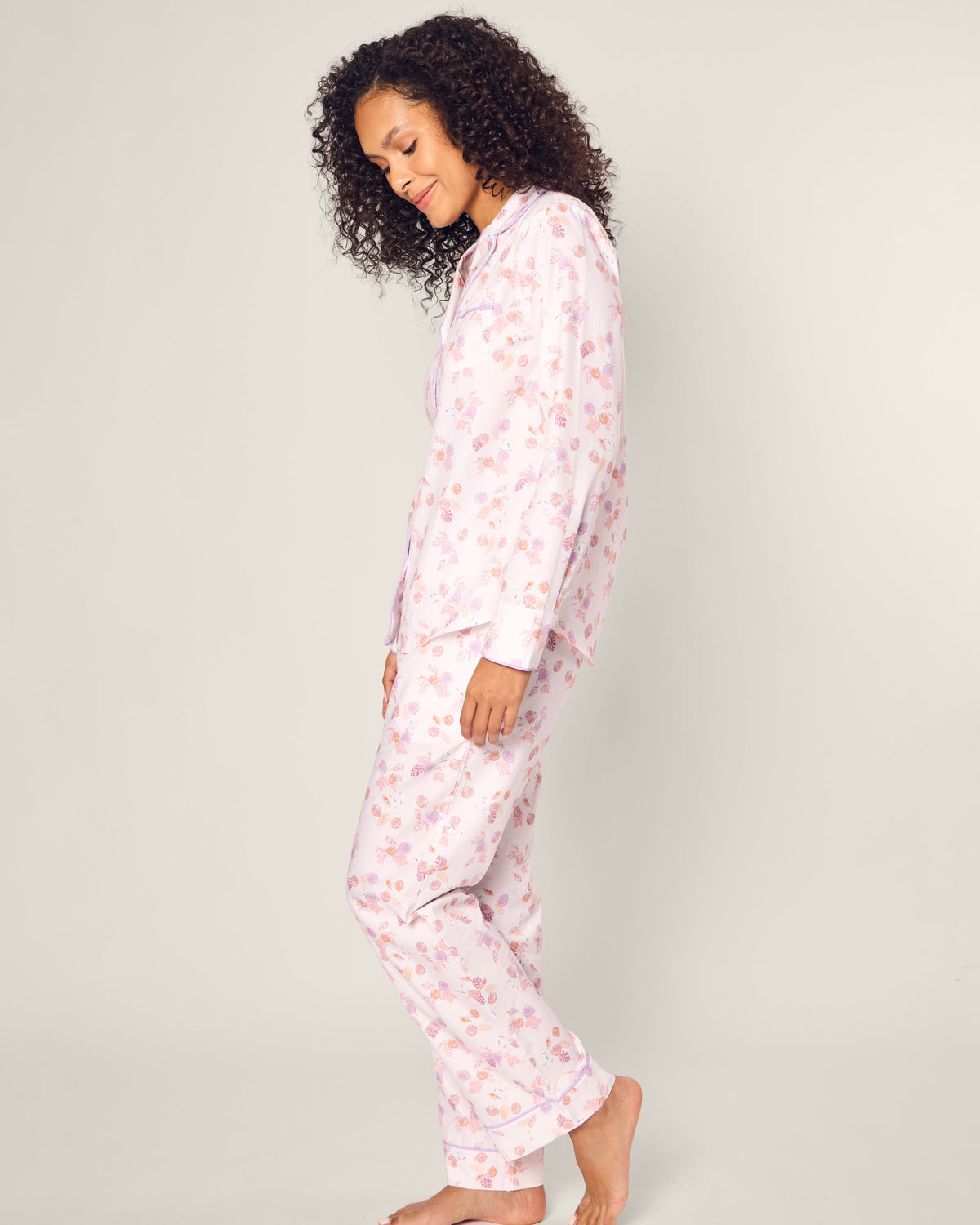 A person with curly hair stands sideways in Petite Plumes Womens Twill Pajama Set in Seashell Splendor. The light pink floral luxury cotton sleepwear contrasts against a plain white backdrop as they smile gently.