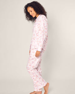 A person with curly hair stands sideways in Petite Plumes Womens Twill Pajama Set in Seashell Splendor. The light pink floral luxury cotton sleepwear contrasts against a plain white backdrop as they smile gently.