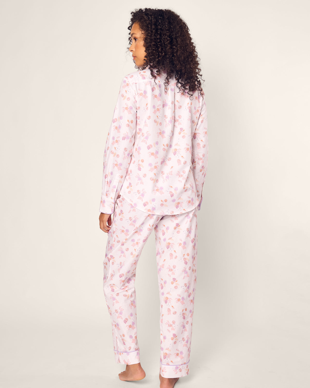 A person with curly hair wears the Petite Plume Womens Twill Pajama Set in Seashell Splendor, made from luxurious cotton sleepwear. It features a soft, comfortable design crafted to perfection against a plain, light background.