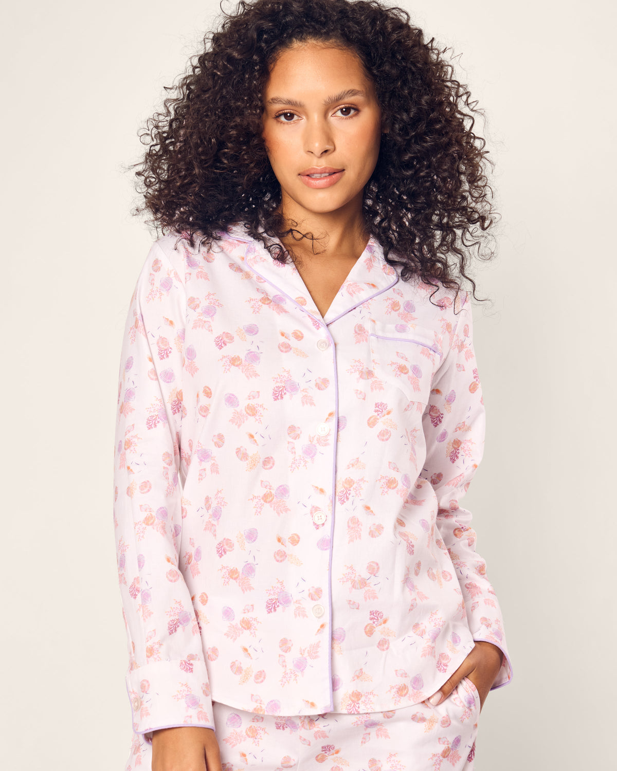 A woman with curly hair models the Petite Plume Womens Twill Pajama Set in Seashell Splendor, featuring a floral pattern on luxurious yarn-dyed fabric. She stands against a white background, slightly tilting her head with one hand by her side.