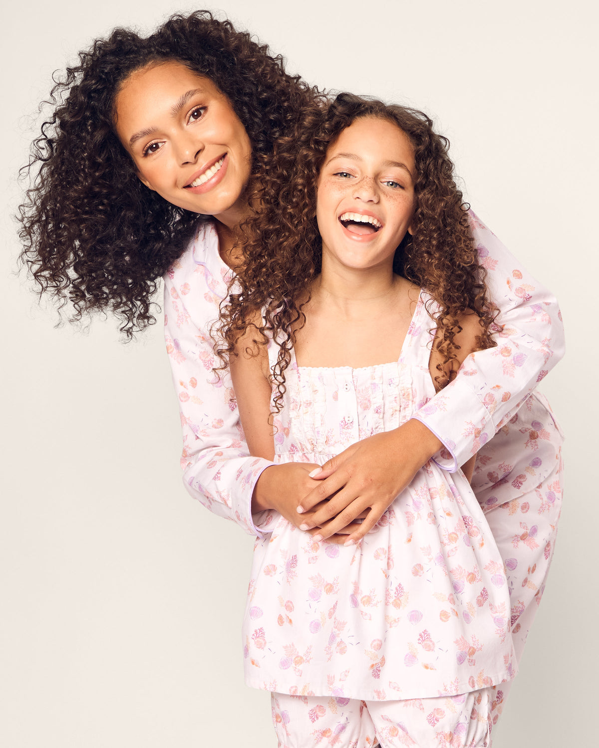 Two curly-haired individuals pose against a plain background, smiling in matching Petite Plume Womens Twill Pajama Sets in Seashell Splendor. The taller person stands behind with arms around the shorter one, who is laughing.