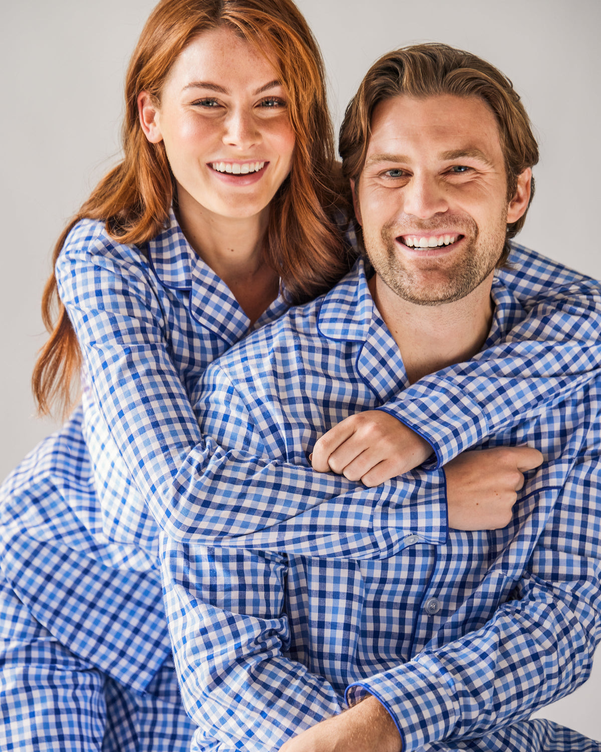 A woman embraces a man as they smile in their matching Petite Plume Womens Twill Pajama Set in Royal Blue Gingham, crafted from quality cotton, both looking cheerful and relaxed against a neutral background.