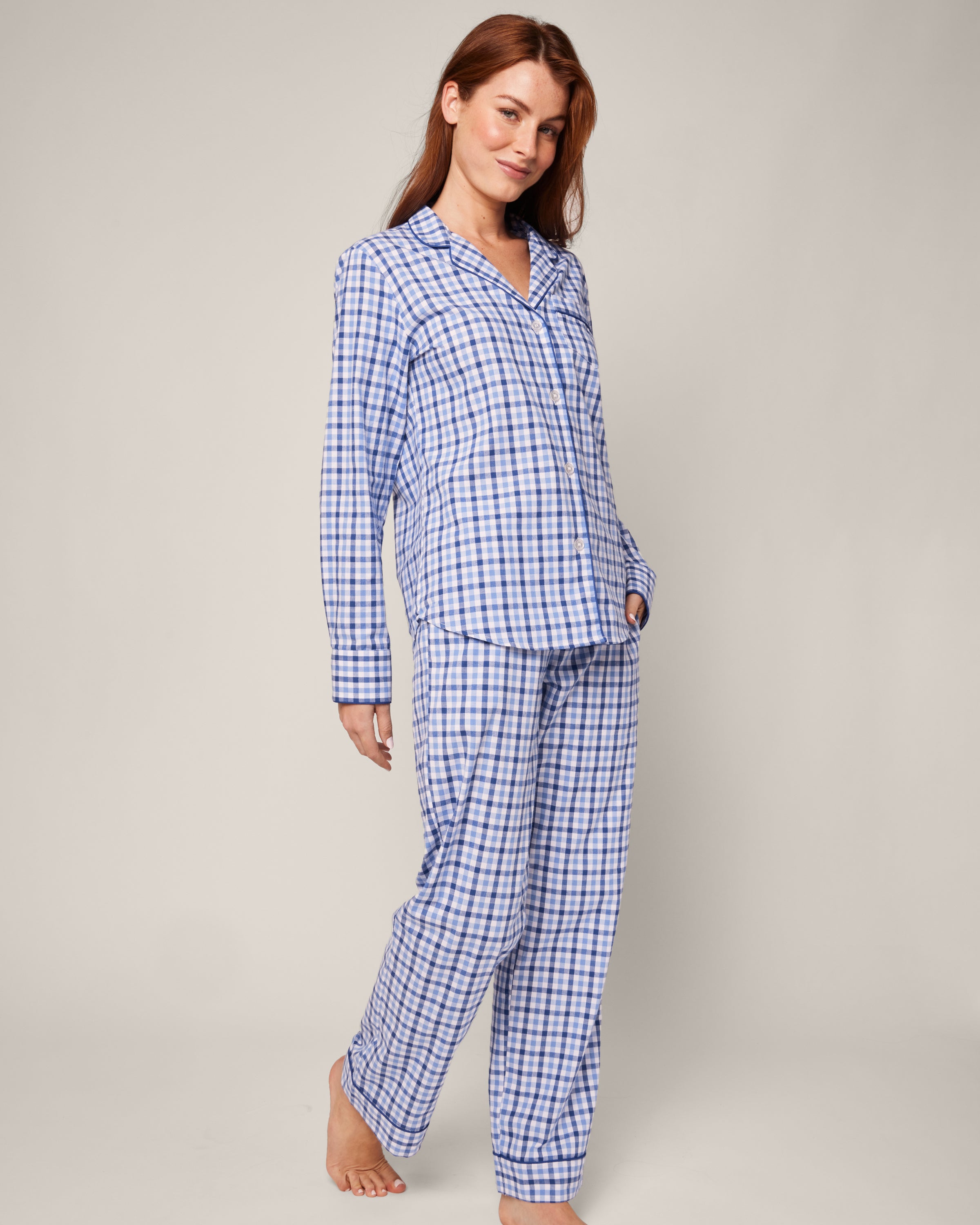 A woman models the Petite Plume Womens Twill Pajama Set in Royal Blue Gingham, standing barefoot against a plain backdrop. With long brown hair and a gentle smile, she places her left hand in her pocket. The chic sleepwear includes a button-up shirt and matching pants, combining comfort and style.