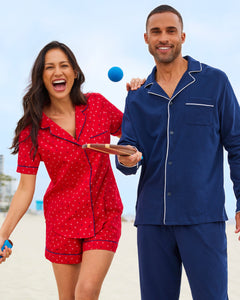 A woman in a red cotton pajama set with white polka dots smiles while holding a ball. Beside her, a man beams at the camera dressed in Petite Plumes Womens Twill Short Sleeve Short Set in Anchors Away, holding a paddle. They seem to be on a sandy beach.