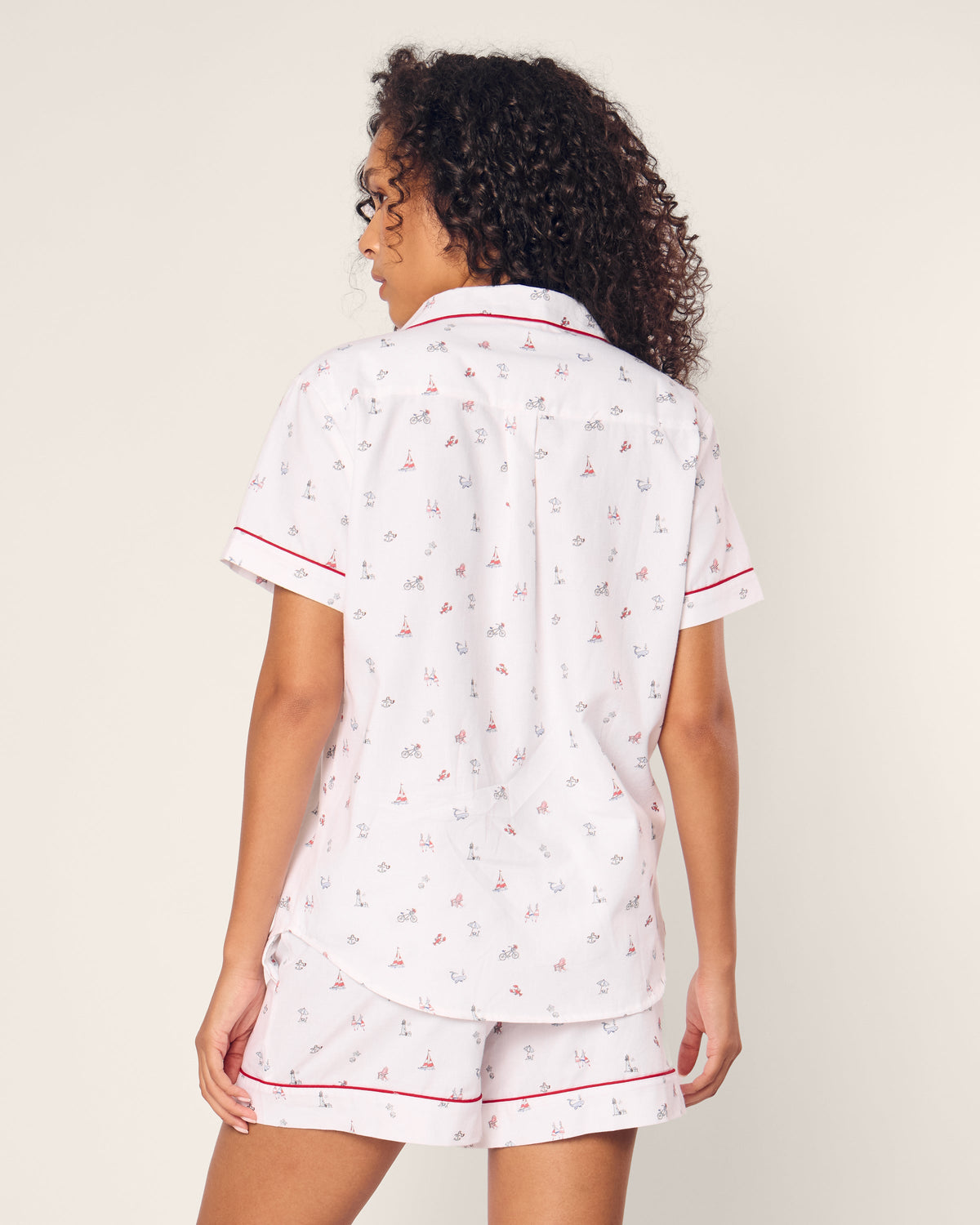 A person with curly hair wearing the Petite Plume Womens Twill Short Sleeve Short Set in Coastal Charms—a luxury cotton sleepwear featuring white fabric with small sailboat designs and red accents—stands out against a plain background.