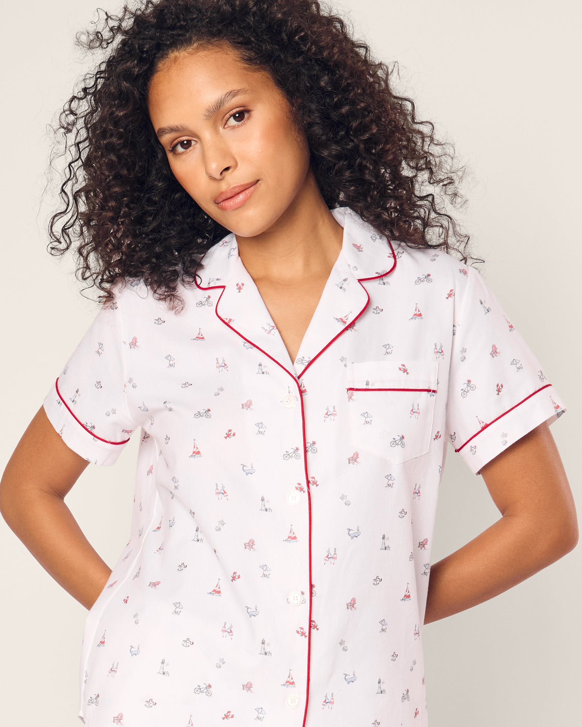 A curly-haired person models the Petite Plume Womens Twill Short Sleeve Short Set in Coastal Charms, featuring premium cotton with vibrant prints and red piping. They stand confidently against a plain background, embodying luxury sleepwear.