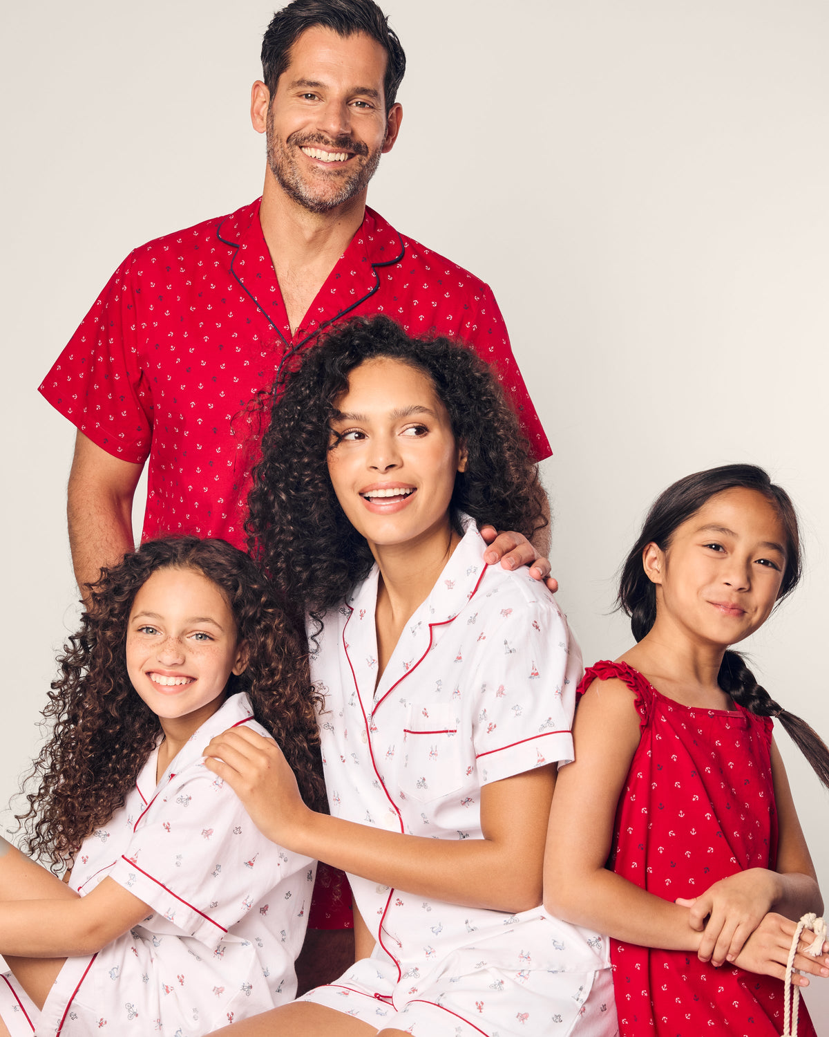 A family poses in matching luxury Petite Plume sleepwear made from the finest quality cotton. The man wears a red shirt while the woman and one girl flaunt white pajamas with red accents. The other girl sports a charming red dress, all smiling warmly.