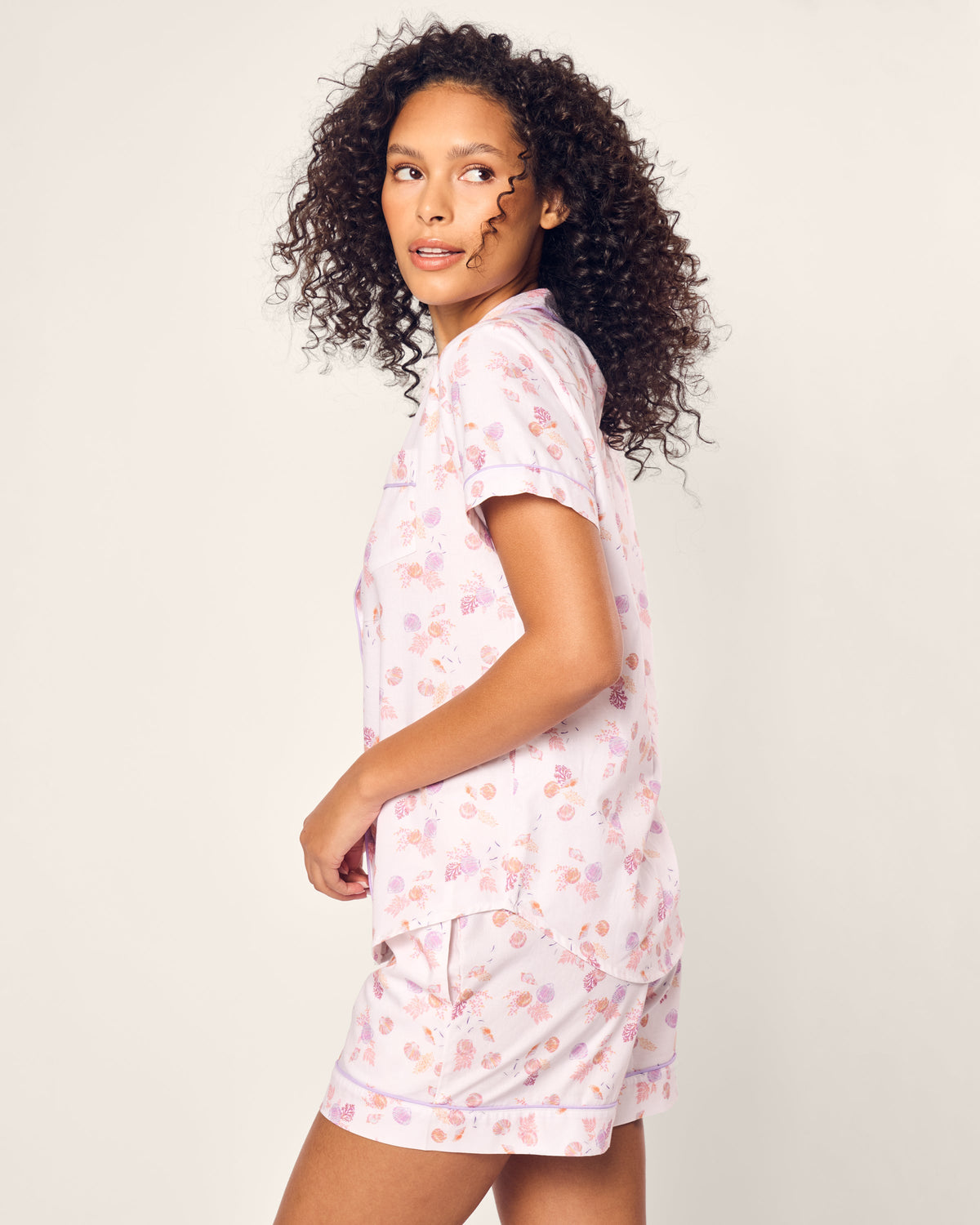 A person with curly hair is wearing Petite Plumes Womens Twill Short Sleeve Short Set in Seashell Splendor, featuring a luxurious floral pattern. Crafted from fine cotton, they stand looking over their shoulder against a neutral background.