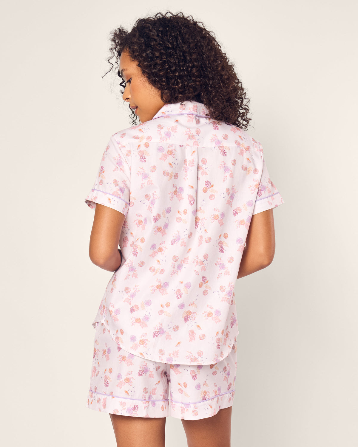 A curly-haired person wears the Womens Twill Short Sleeve Short Set in Seashell Splendor by Petite Plume. This luxurious pajama set features a light pink floral pattern, crafted from premium cotton, with a short-sleeve shirt and shorts. They exude comfort and style against a plain background.
