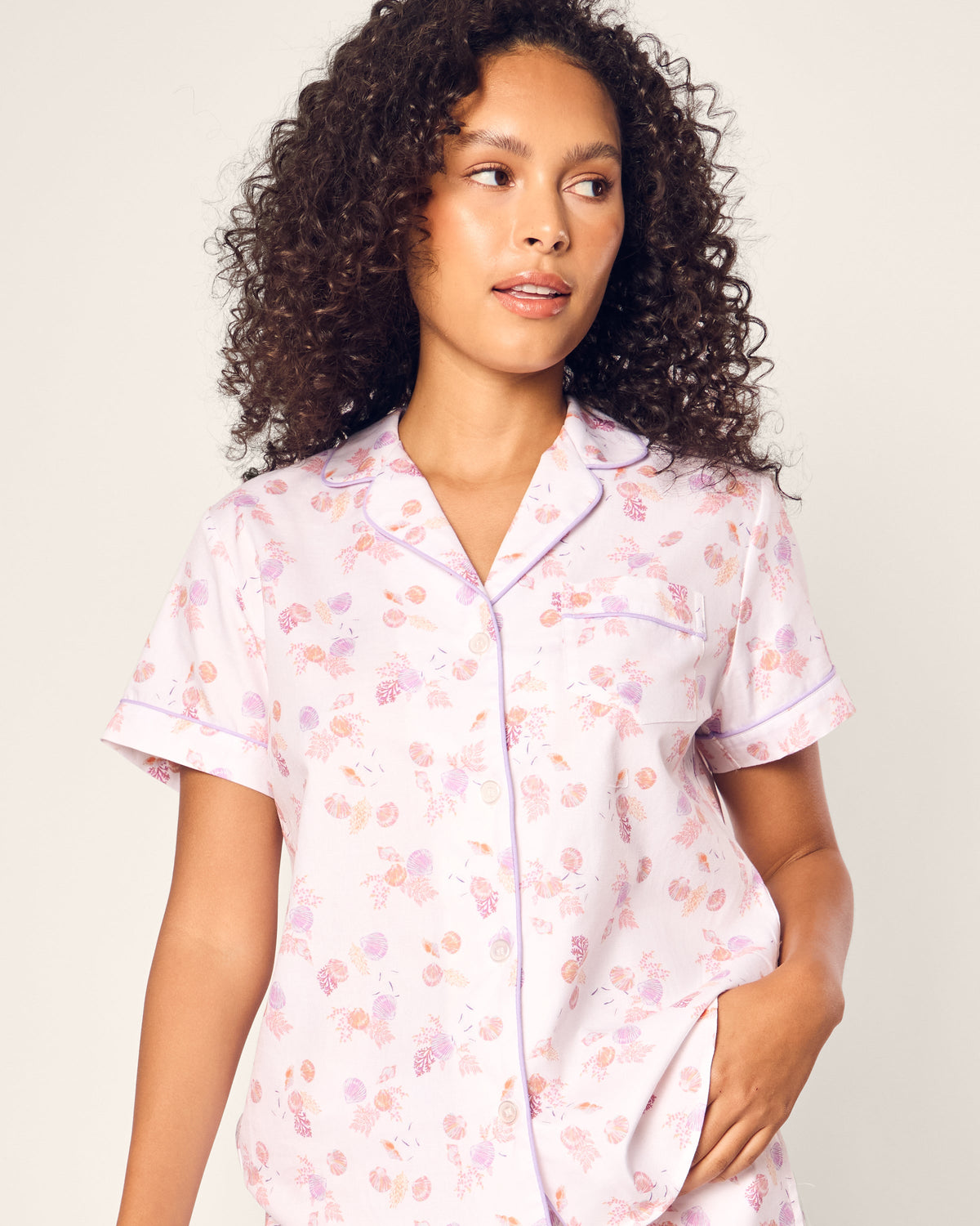 A person with curly hair wears the Womens Twill Short Sleeve Short Set in Seashell Splendor by Petite Plume, looking to the side neutrally. The light-colored set features pink and orange floral patterns, crafted from high-quality cotton, with short sleeves and a buttoned front.
