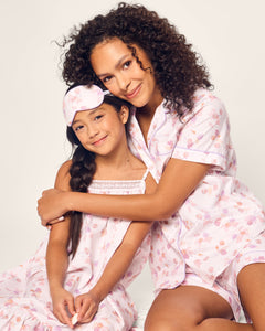 A woman and girl in matching Petite Plume Seashell Splendor short sleeve pajamas—a luxe cotton twill set—smile and embrace. The girls braided hair and eye mask complete their charming look against a light, neutral backdrop.