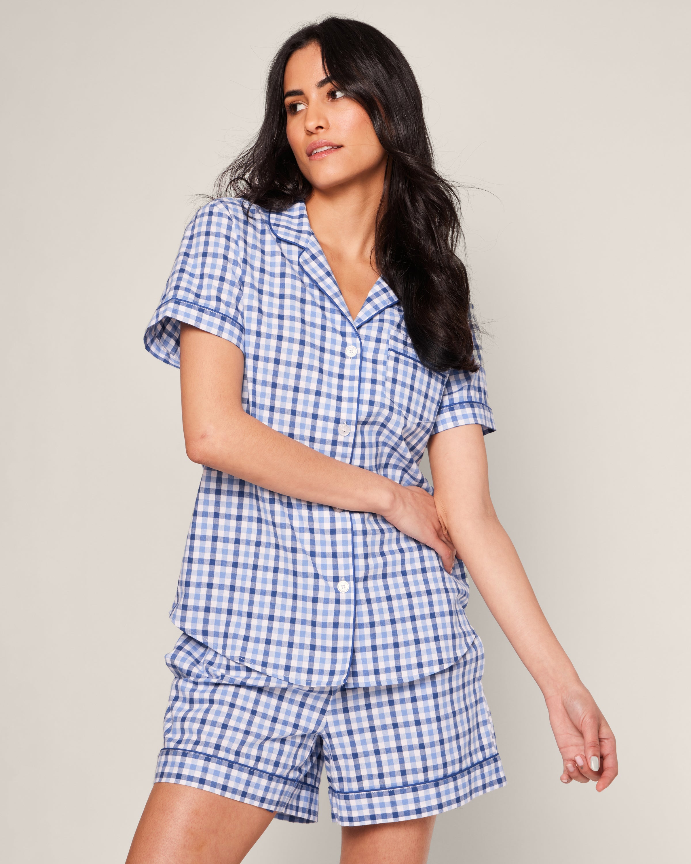 A woman in a Petite Plume Royal Blue Gingham Twill Pajama Short Set stands against a plain backdrop. With her long, dark hair slightly turned to the side and gently holding one arm, she exudes luxury sleepwear made from the finest quality cotton.