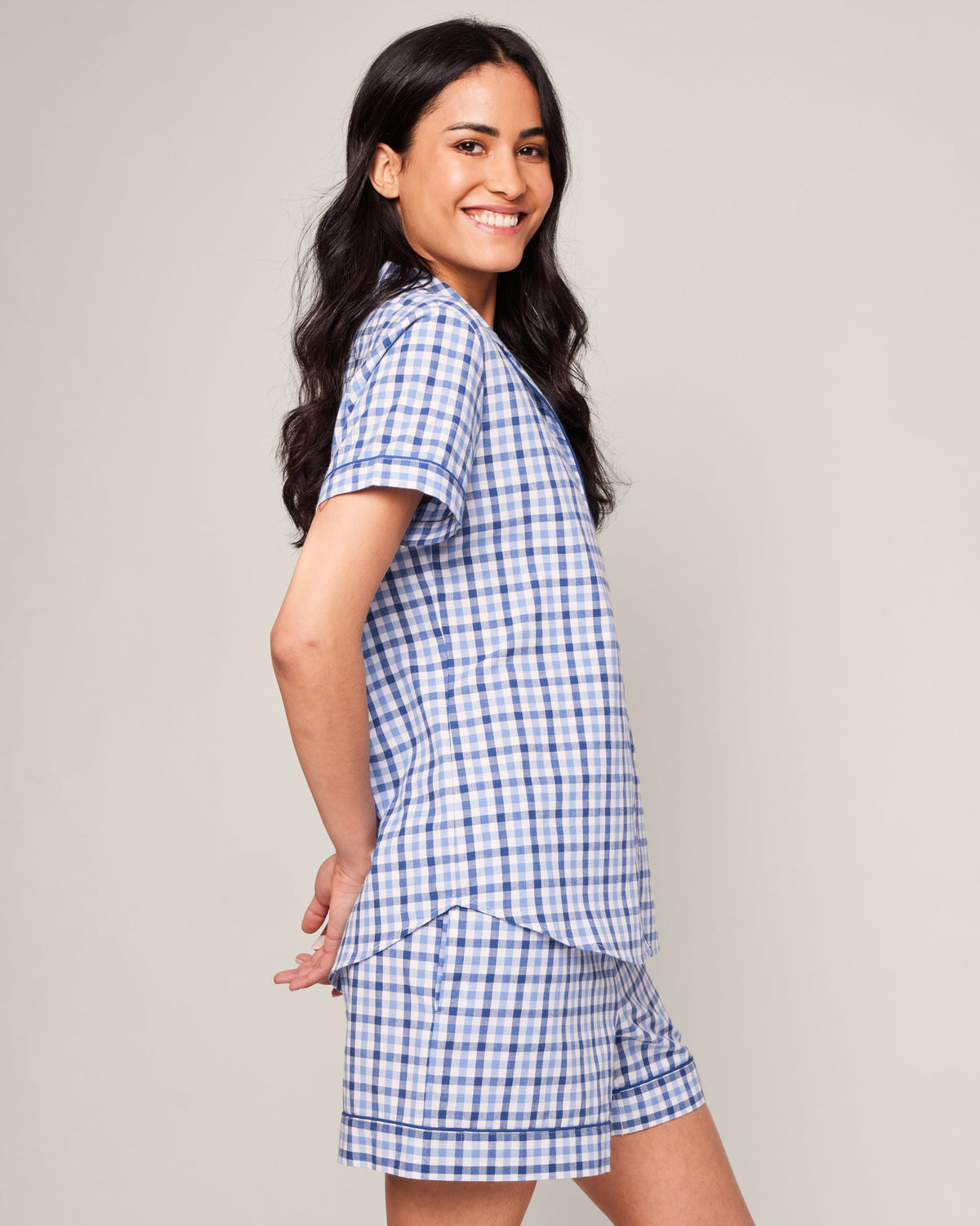 A woman with long dark hair smiles while wearing the Petite Plume Womens Twill Pajama Short Sleeve Short Set in Royal Blue Gingham. She stands sideways, hands behind her back, against a plain light background.