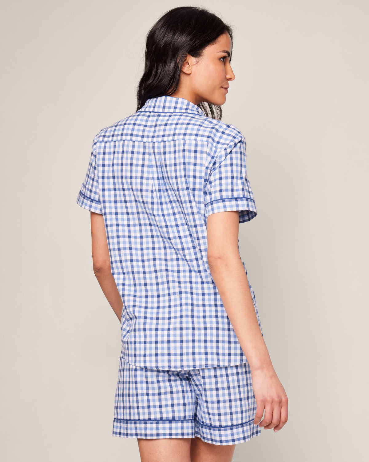 A person with long dark hair is wearing the Womens Twill Pajama Short Sleeve Short Set in Royal Blue Gingham by Petite Plume, standing sideways against a neutral background.