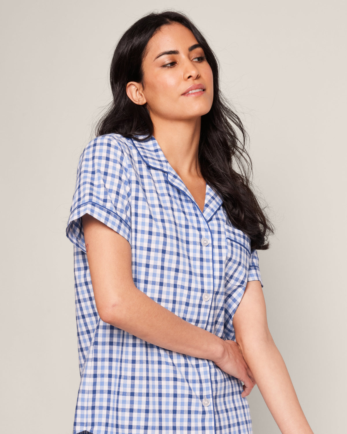 A woman with long dark hair wears the Womens Twill Pajama Short Sleeve Short Set in Royal Blue Gingham by Petite Plume. She gazes serenely to the side, enveloped in high-quality cotton, against a plain light background that highlights her tranquil aura.
