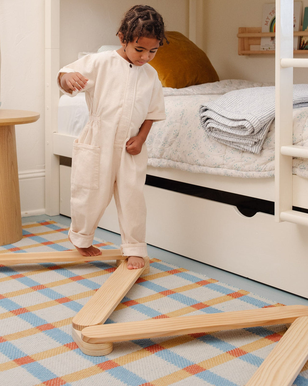 Toddler balancing barefoot on wooden Acrobat balance beams arranged on a checkered rug in a bedroom