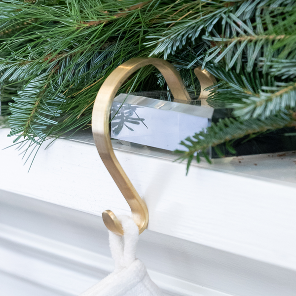 Close-up of acrylic stocking holder with brass hook showing clear acrylic block resting on white mantel with evergreen garland