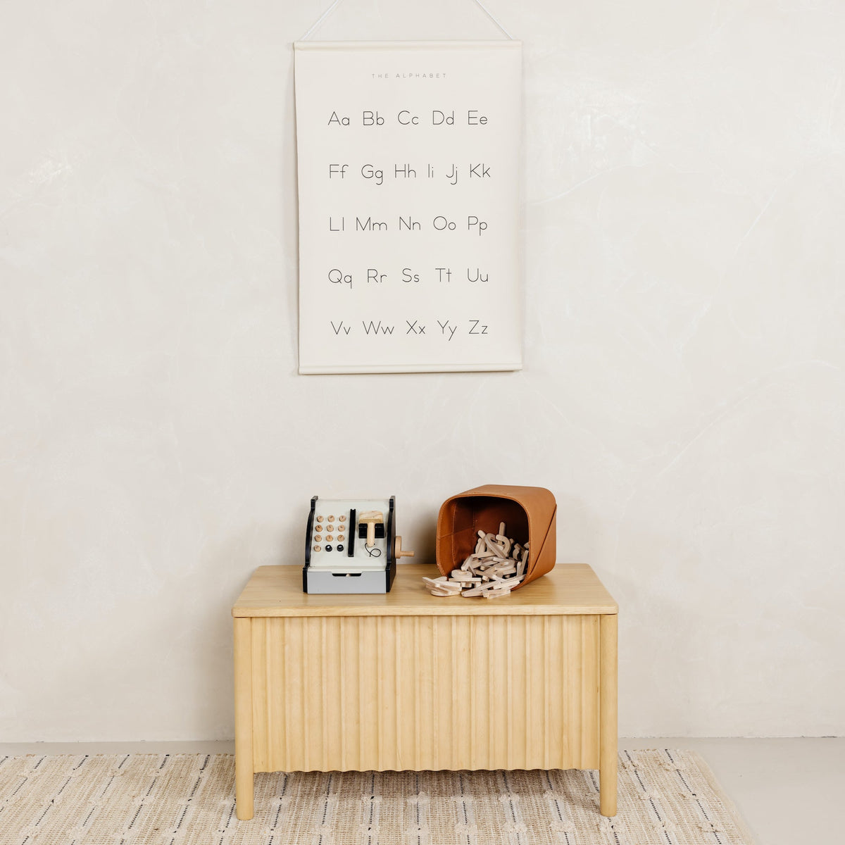 Alphabet poster labeled The Alphabet hanging on a pale wall above a wooden bench with a toy cash register and a tipped leather basket of wooden toys