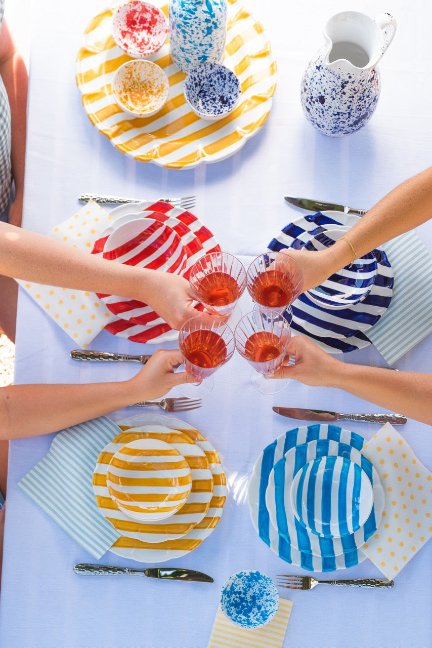 Four people toasting above a table set with Amalfitana striped bowls and plates in red, blue, yellow, and aqua
