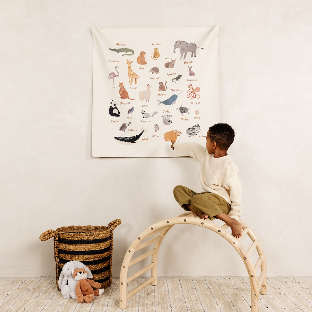 Child sitting on a wooden climbing arch pointing at a hanging animal alphabet tapestry in a neutral nursery setting