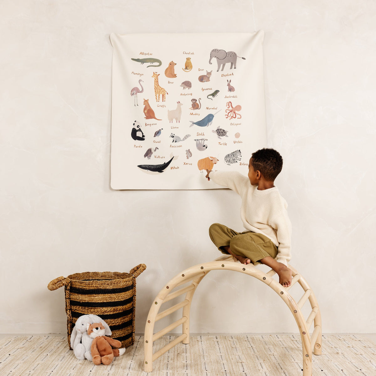 Child sitting on a wooden climbing arch pointing at a hanging animal alphabet tapestry in a neutral nursery setting
