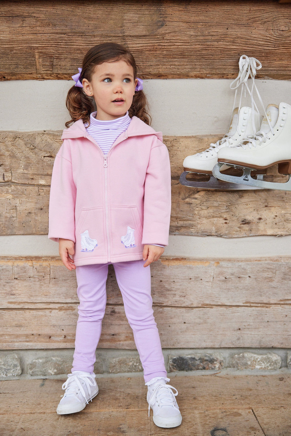 Little girl wearing lavender-striped turtleneck with pink hoodie and leggings, standing by wood wall with hanging ice skates