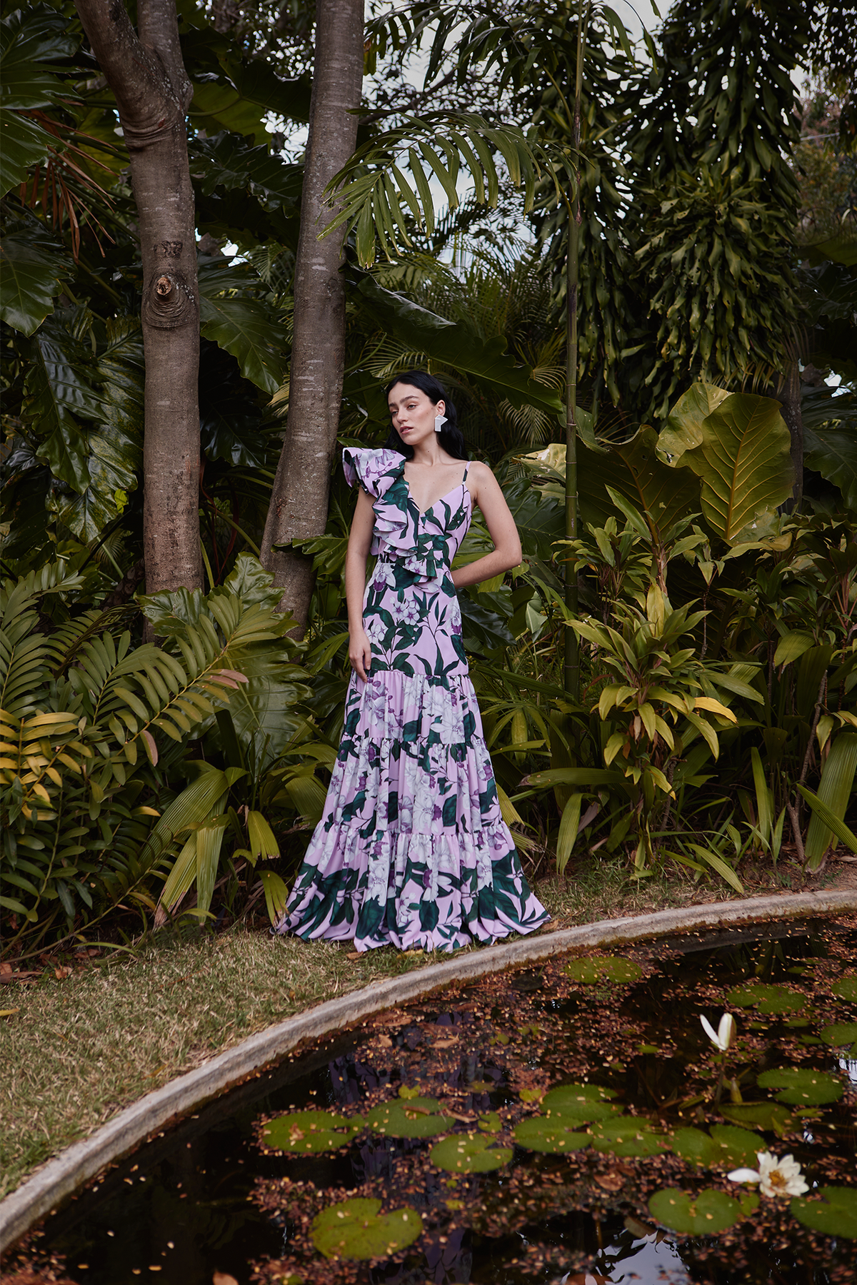 Model wearing lavender Aracea maxi dress with ruffled one-shoulder and tiered skirt beside a lily pond in a tropical garden