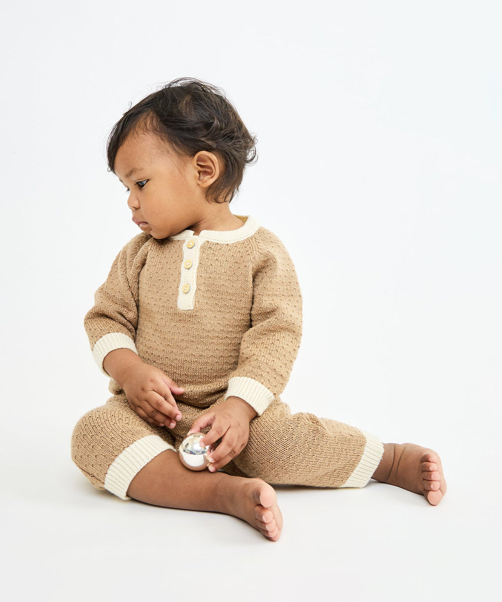 Seated front-side view of Archie Jumpsuit in camel on baby showing knit texture, cream contrast trim and button placket on white background