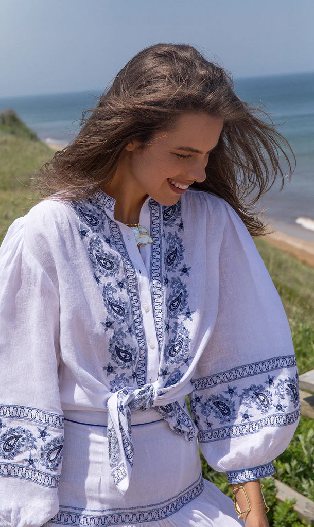 Front view of Seaside Blaire Blouse showcasing blue paisley embroidery, tie-front, billowy linen sleeves with beach background